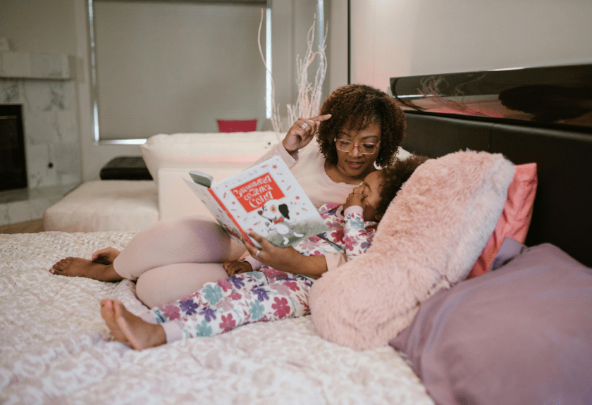 Black woman lies on bed with young child.They read a book together.